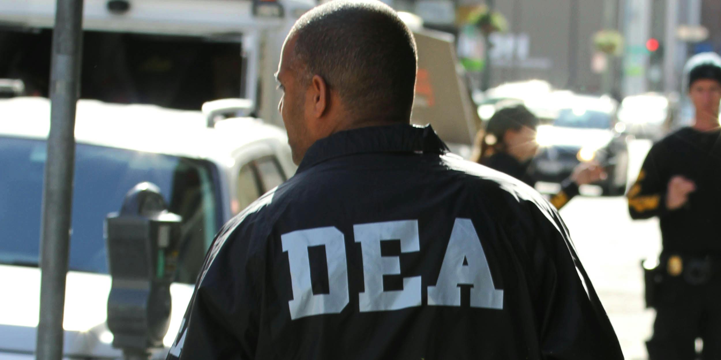 DEA agent stands with back to camera, DEA white lettering visible on jacket