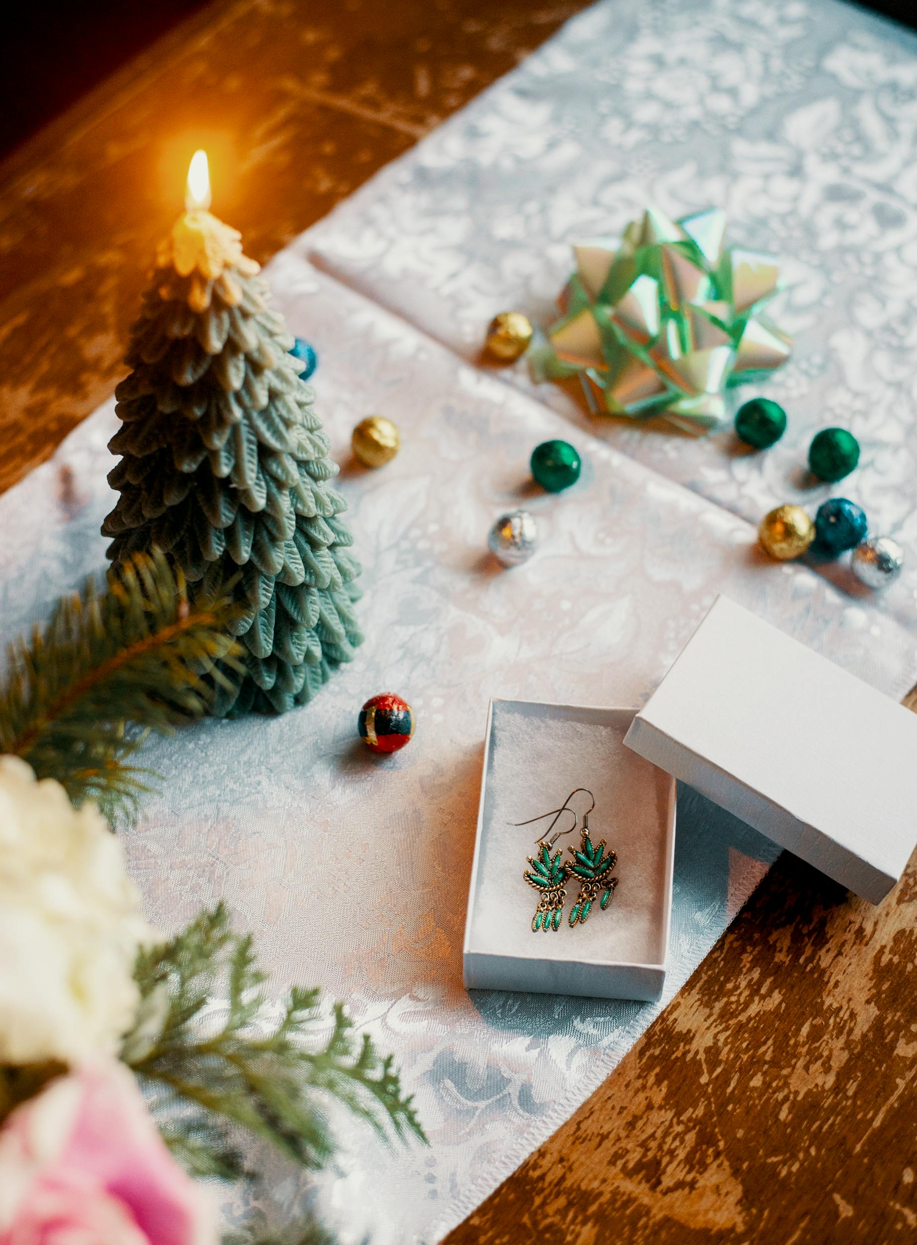 A decorative spread with a Christmas tree candle, several baubles and a green bow. There is a box with two weed leaf shaped earrings