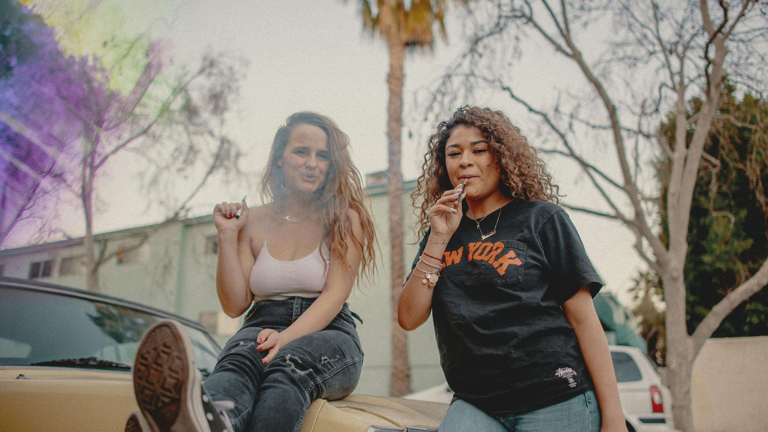 Two women sit on the hood of a car and share the best weed pen