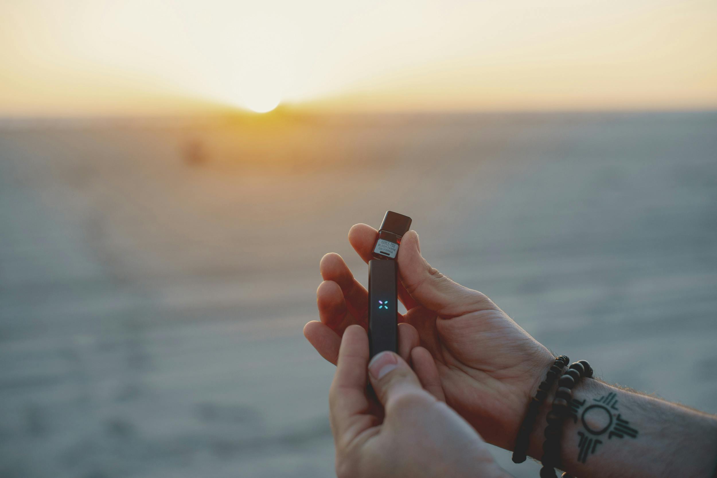 Hands holding one of the best weed vaporizers, the PAX ERA, in front of a sunset on the beach.