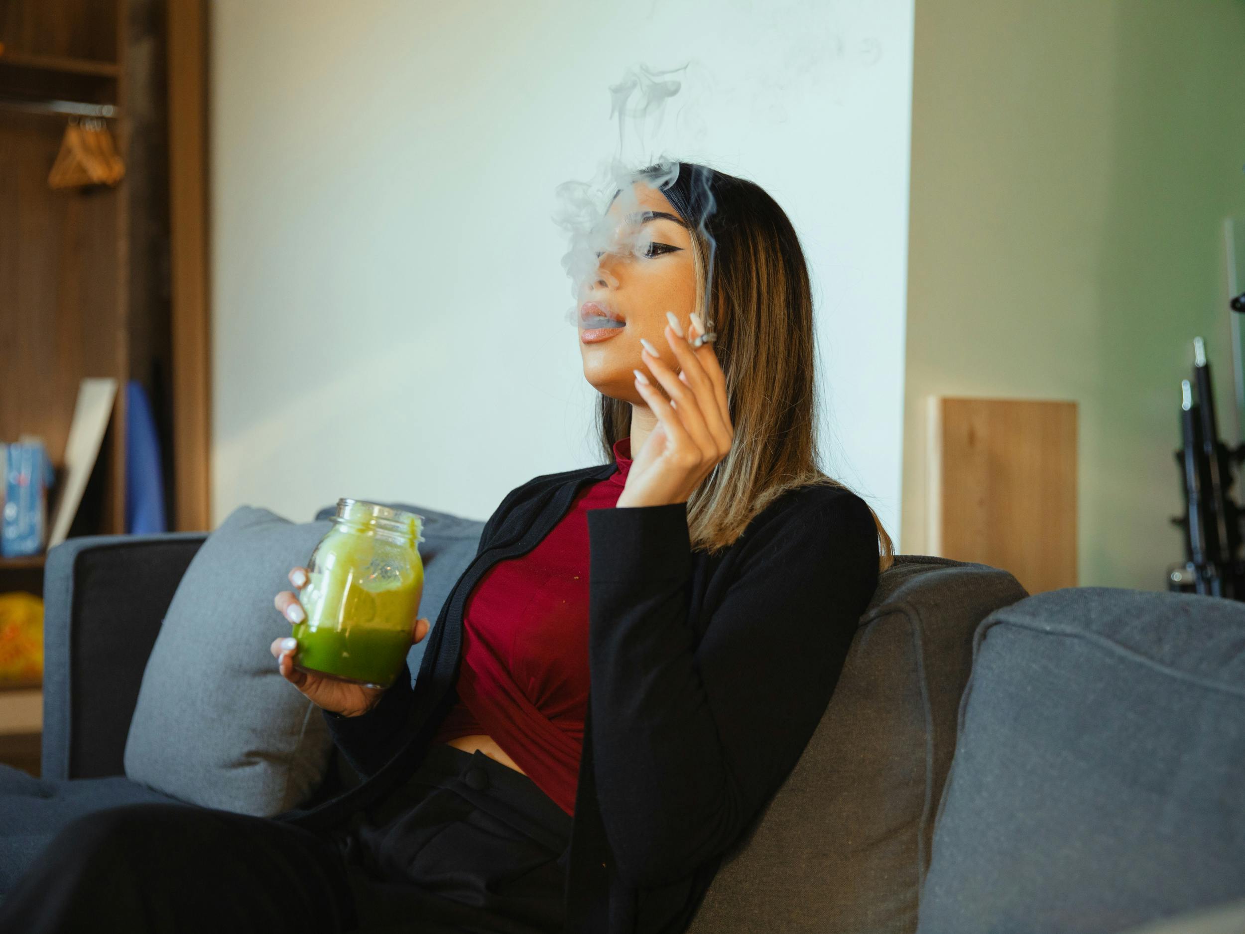 A woman sits on a couch smoking a joint and drinking a mason jar full of Bhang. Learn how to make bhang at home with this guide.