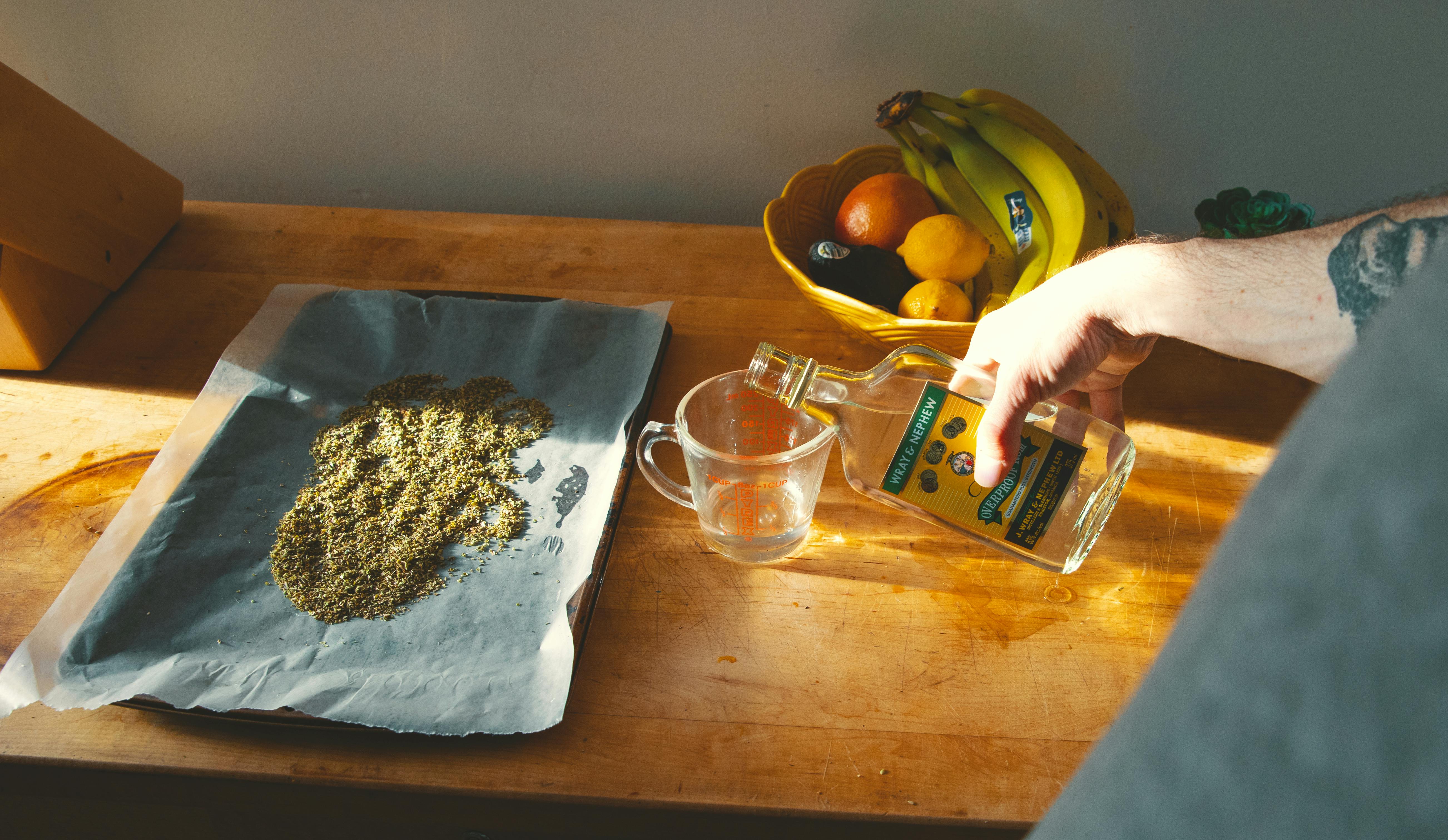 A hand pours alcohol into a measuring cup next to a baking sheet covered in ground up flower. Learn how to make cannabis tincture with this handy guide.